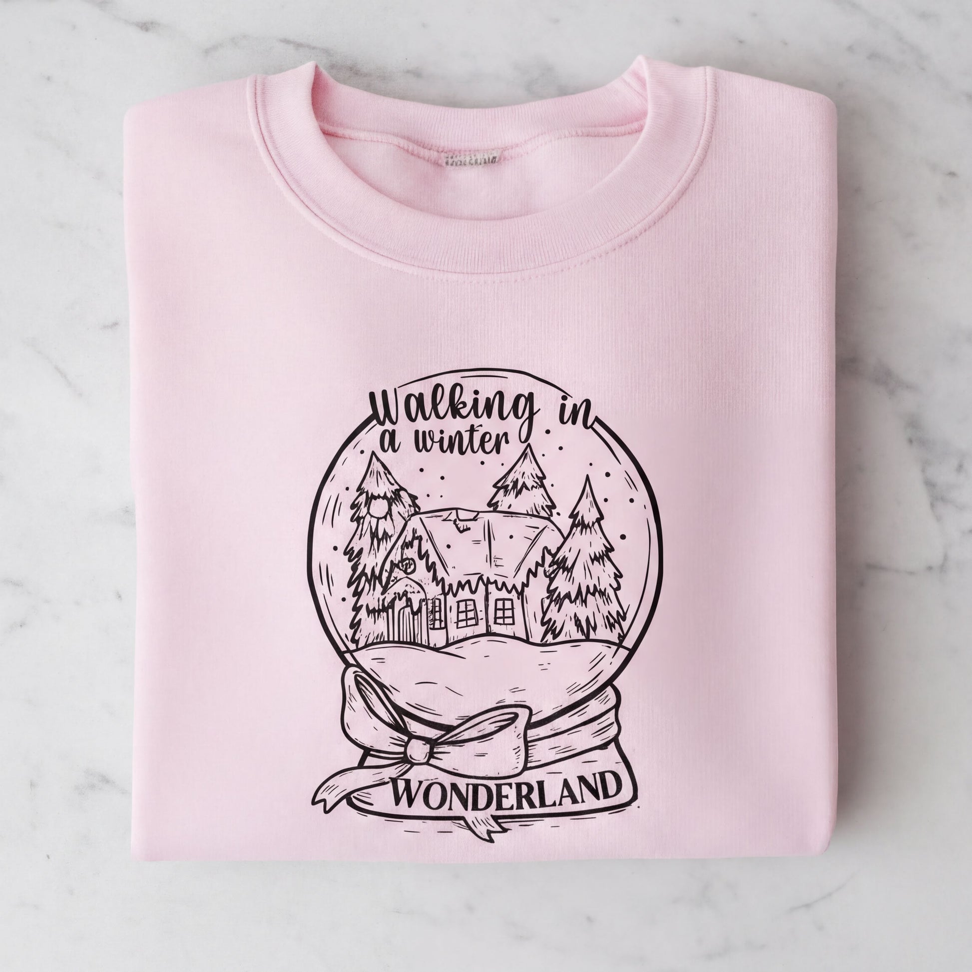 A pink sweatshirt with a graphic design depicting a snow globe containing a winter scene with a house and trees. The text on the sweatshirt reads "Walking in winter wonderland".