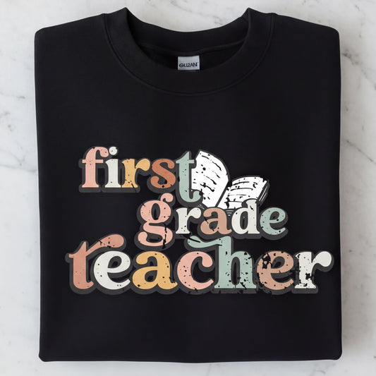 A black sweatshirt with the text "first grade teacher" printed on it in a colorful, distressed font.