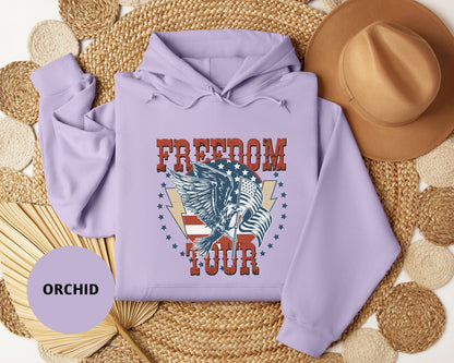 A purple hoodie with a graphic design of a winged eagle and the text "FREEDOM TOUR" is displayed on a woven mat, along with a brown hat and a fan.