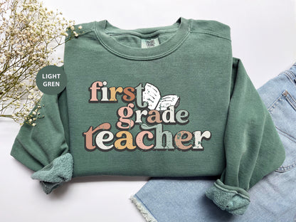 A green sweatshirt with the text "first grade teacher" printed on it, along with a butterfly graphic. The sweatshirt is laid out on a white surface, with some denim jeans visible in the background.