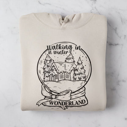 A beige hoodie with a black and white illustration of a snow globe depicting a winter scene, along with the text "Walking in winter wonderland" printed on it.
