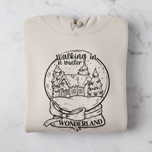 A beige hoodie with a black and white illustration of a snow globe depicting a winter scene, along with the text "Walking in winter wonderland" printed on it.
