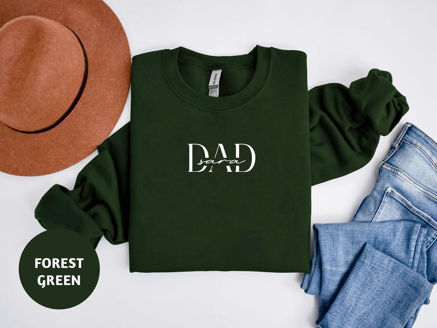 A green sweatshirt with the text "DAD" printed on it, along with a brown hat and a pair of blue jeans, all laid out on a white background.