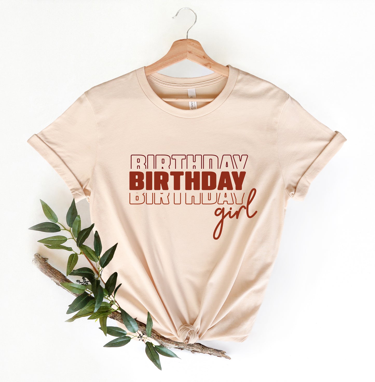 A beige t-shirt with the text "BIRTHDAY BIRTHDAY girl" printed on it, along with a wooden hanger.