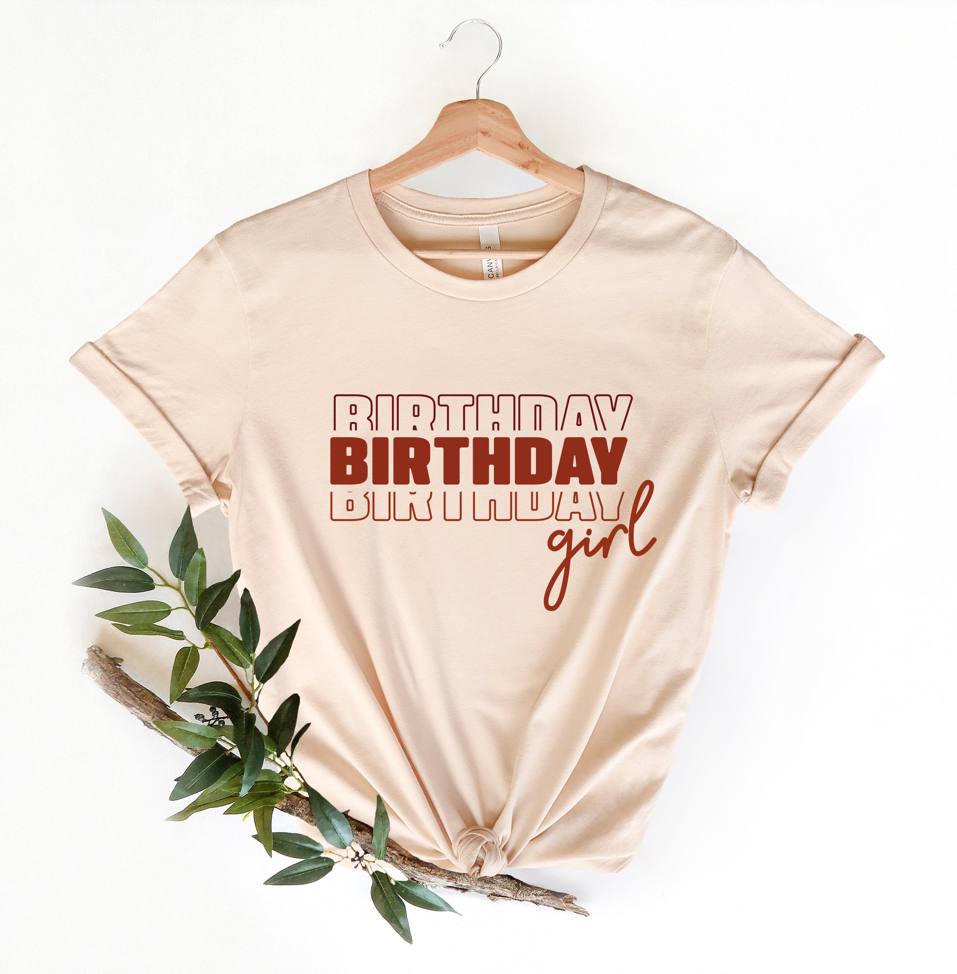 A beige t-shirt with the text "BIRTHDAY BIRTHDAY girl" printed on it, along with a wooden hanger.