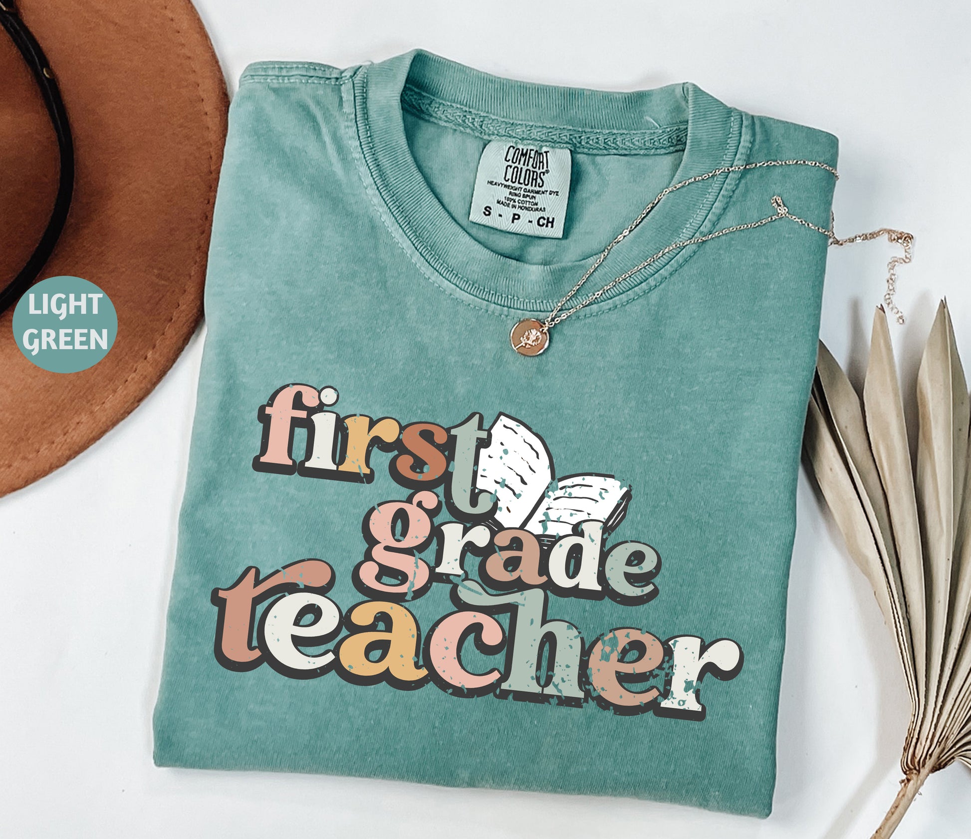 A turquoise t-shirt with the text "first grade teacher" printed on it, along with a necklace and a brown hat.