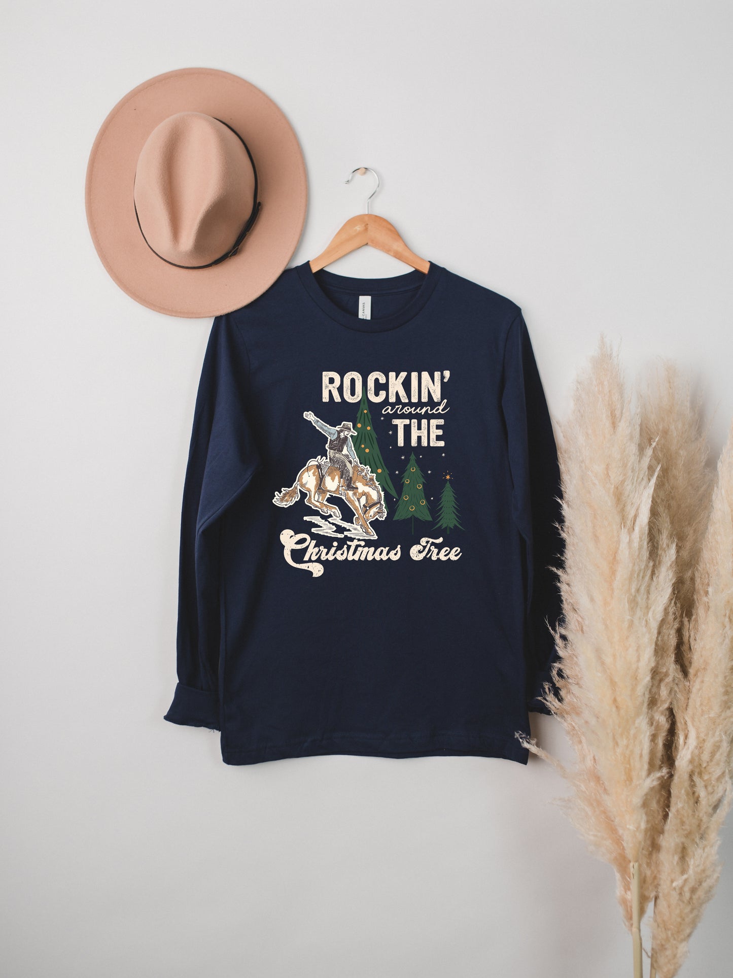 A navy blue long-sleeved shirt with a graphic design and text, hanging on a wooden hanger. The shirt features a cowboy riding a bucking bronco, with the text "ROCKIN' around THE CHRISTMAS Tree".