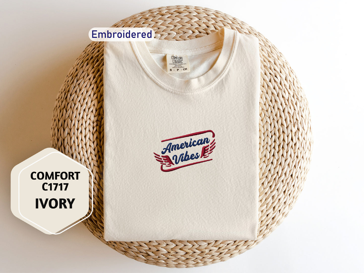 a white t-shirt with a red and blue logo, laid out on a woven mat. The t-shirt appears to be embroidered, and there is a label on the collar. The image also includes text that reads "Embroidered" and "COMFORT C1717 IVORY".