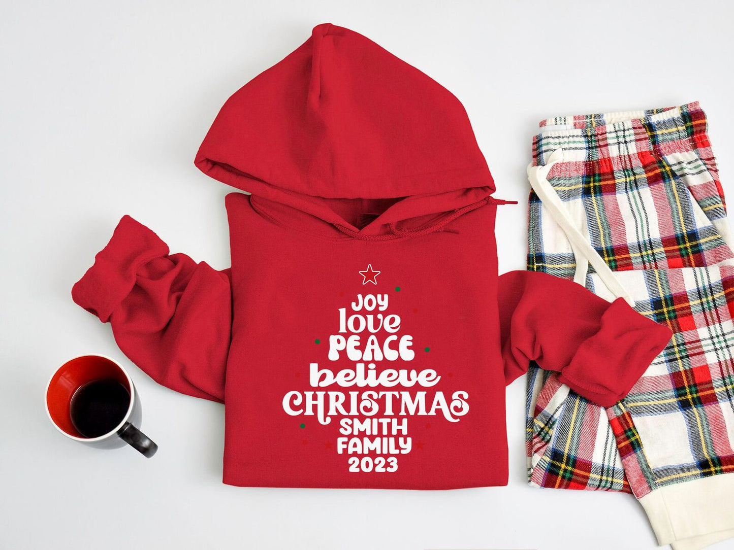 A red hoodie with a Christmas tree design and text, a pair of plaid pajama pants, and a mug on a white background.