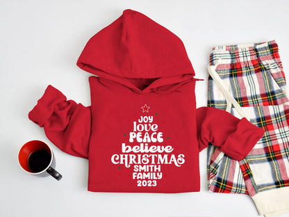 A red hoodie with a Christmas tree design and text, a pair of plaid pajama pants, and a mug on a white background.