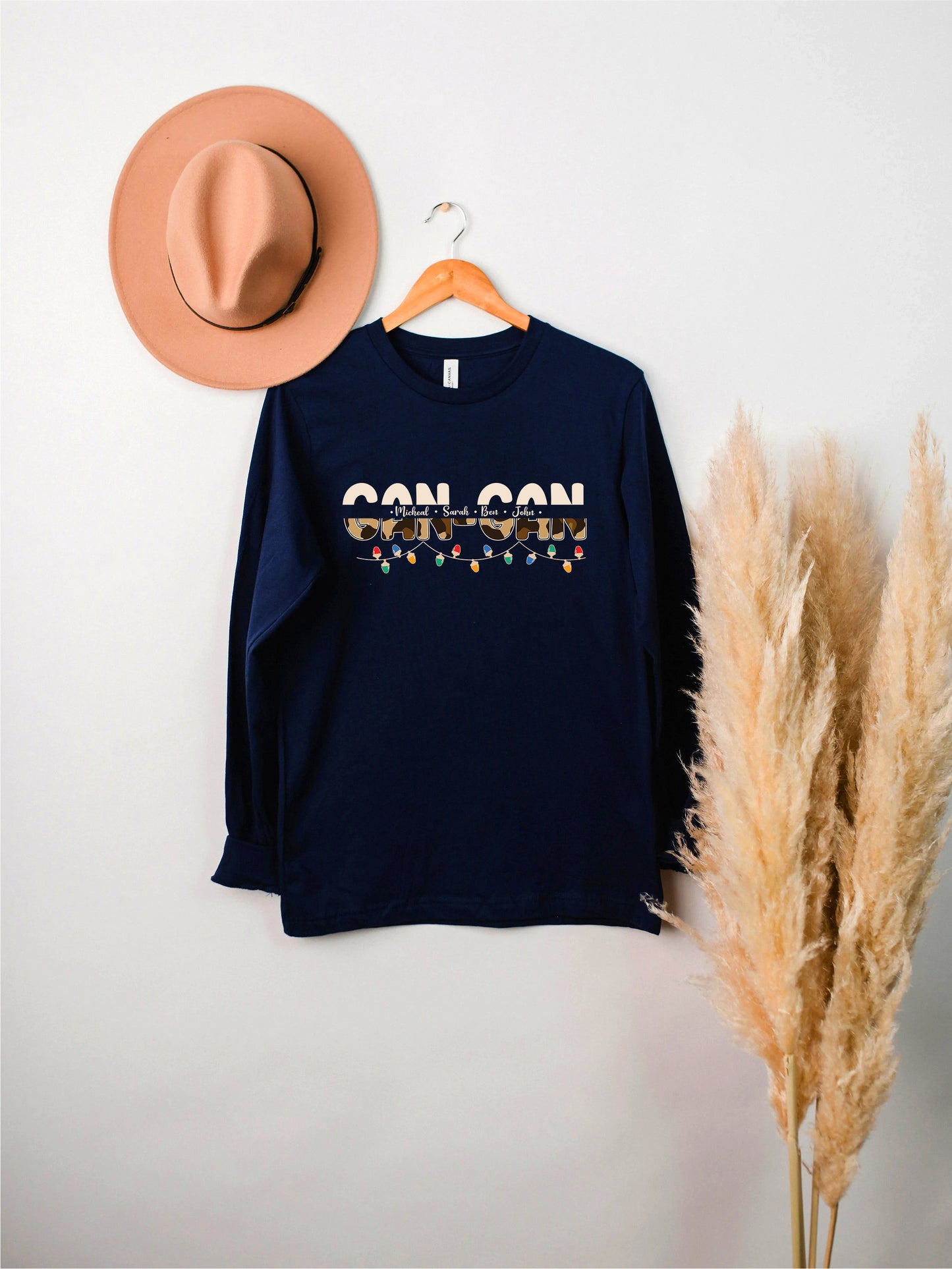 a navy blue long-sleeved shirt with the word "CAN CON" printed on it, hanging on a wooden hanger. Next to the shirt, there is a pink hat and some potted plants, creating a casual and stylish setting.