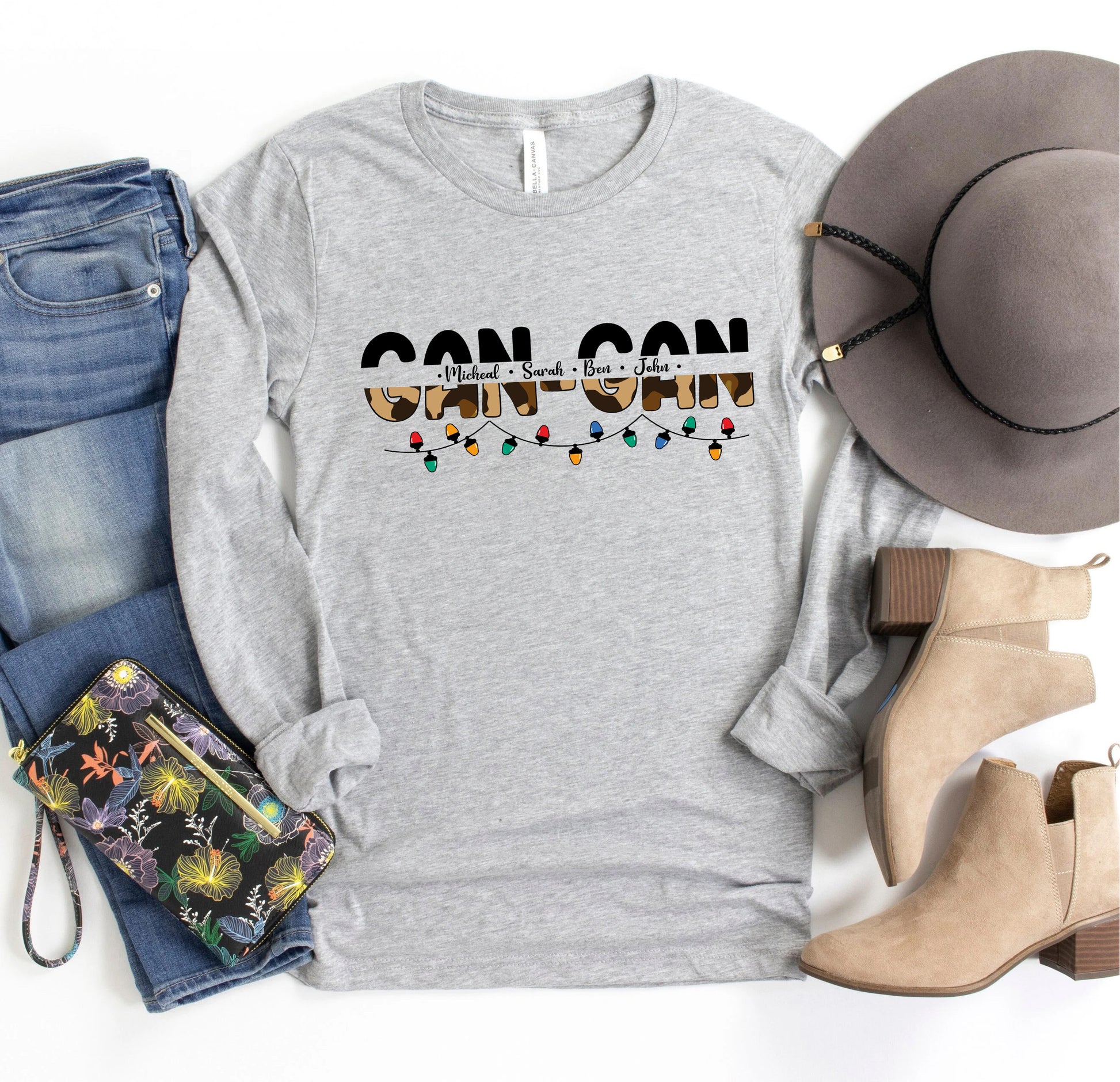 A casual outfit consisting of a gray long-sleeved t-shirt with a graphic design, a pair of blue jeans, a gray hat, and beige ankle boots, along with a floral-patterned purse and a pair of sunglasses.