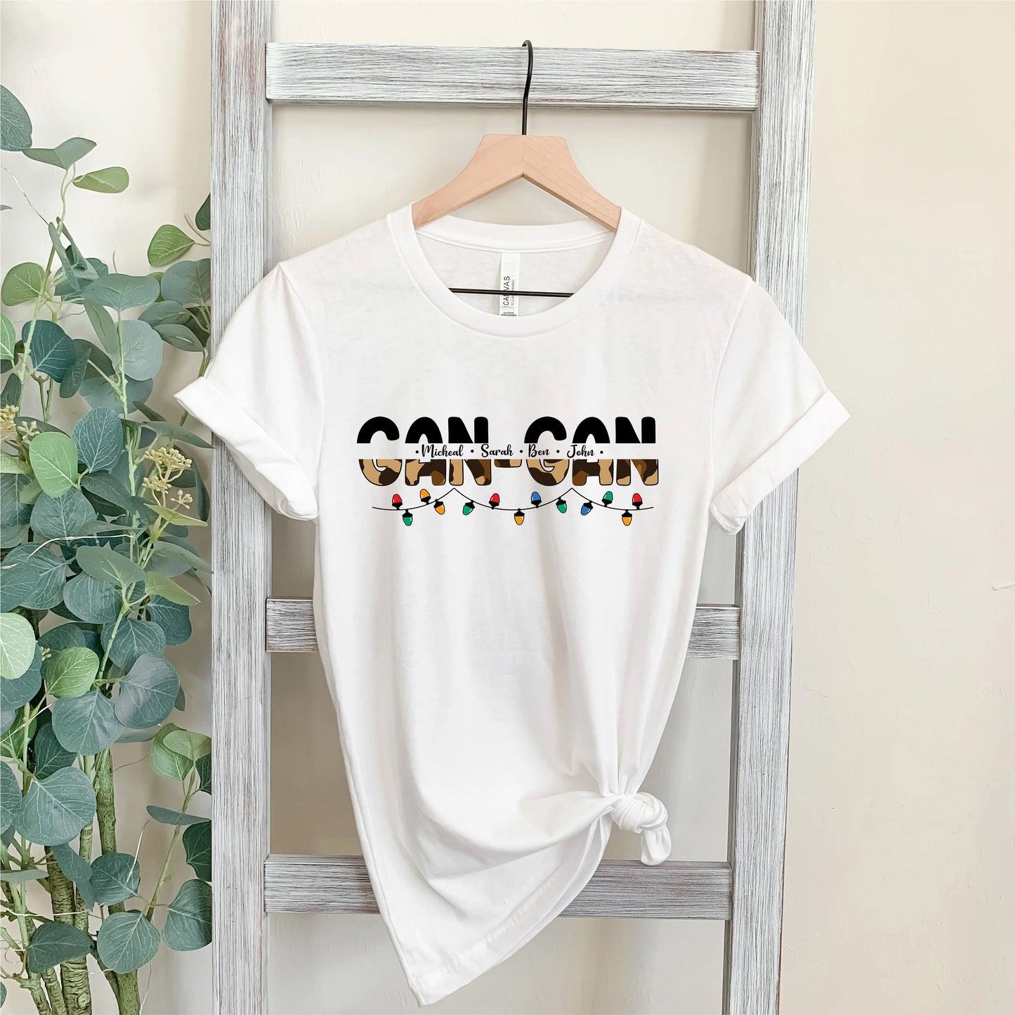 A white t-shirt with the text "CAN CAN" printed on it, hanging on a wooden hanger.
