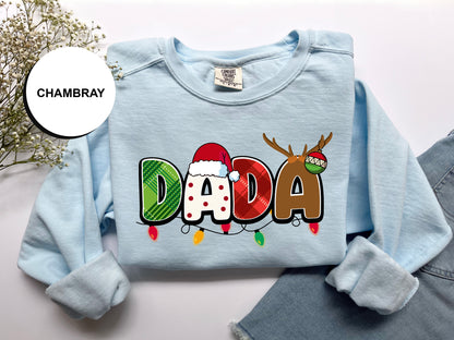 A light blue long-sleeved shirt with the word "DADA" printed in large letters, along with a Santa hat and antlers, is displayed on a white background.