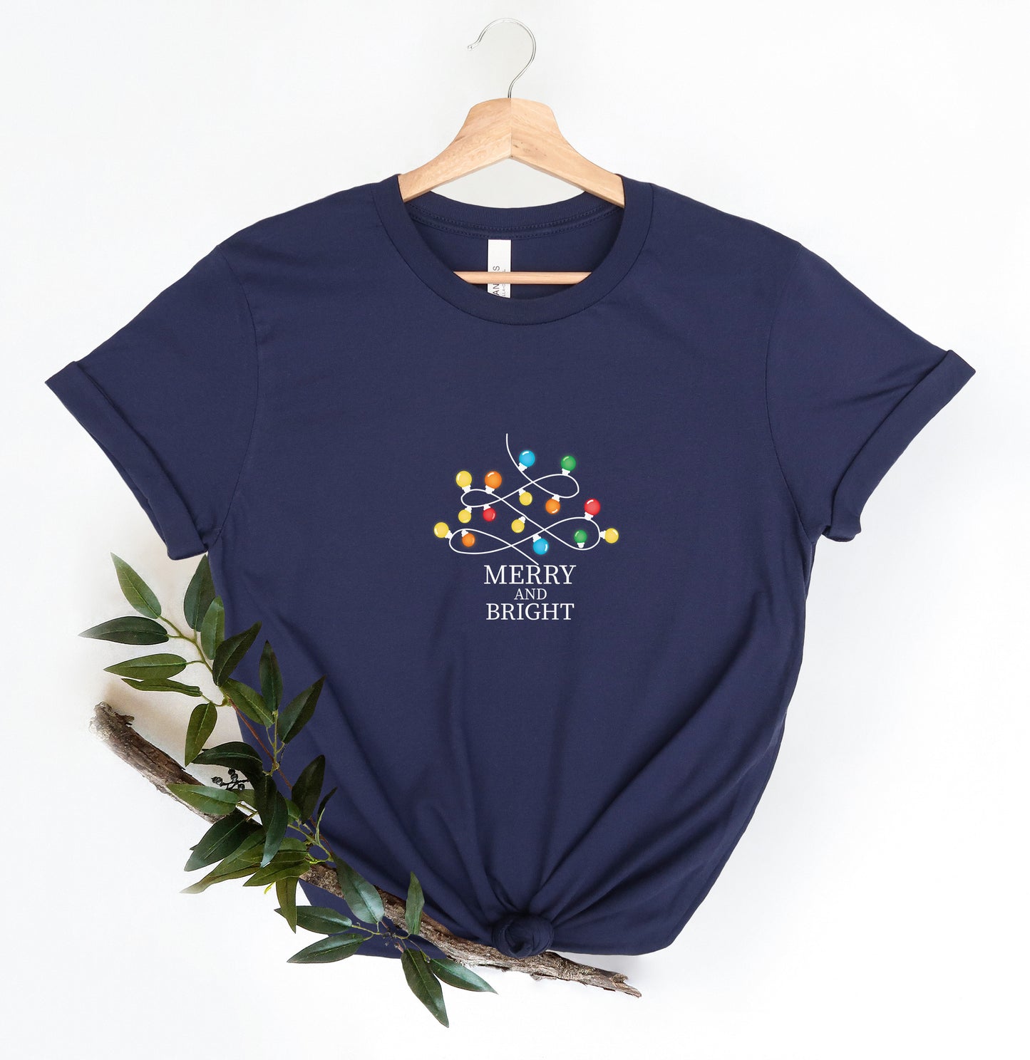 A navy blue t-shirt with a graphic design of colorful lights and the text "MERRY IS BRIGHT" printed on it, hanging on a wooden hanger.