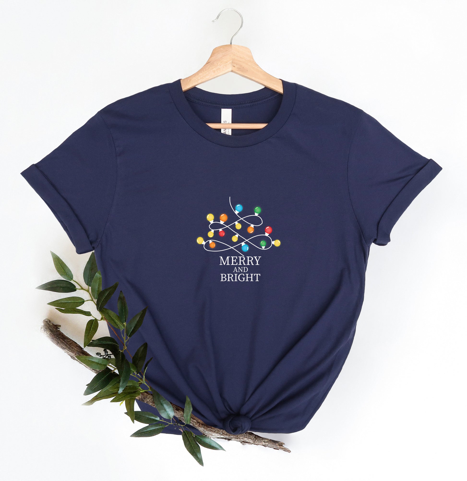 A navy blue t-shirt with a graphic design of colorful lights and the text "MERRY IS BRIGHT" printed on it, hanging on a wooden hanger.