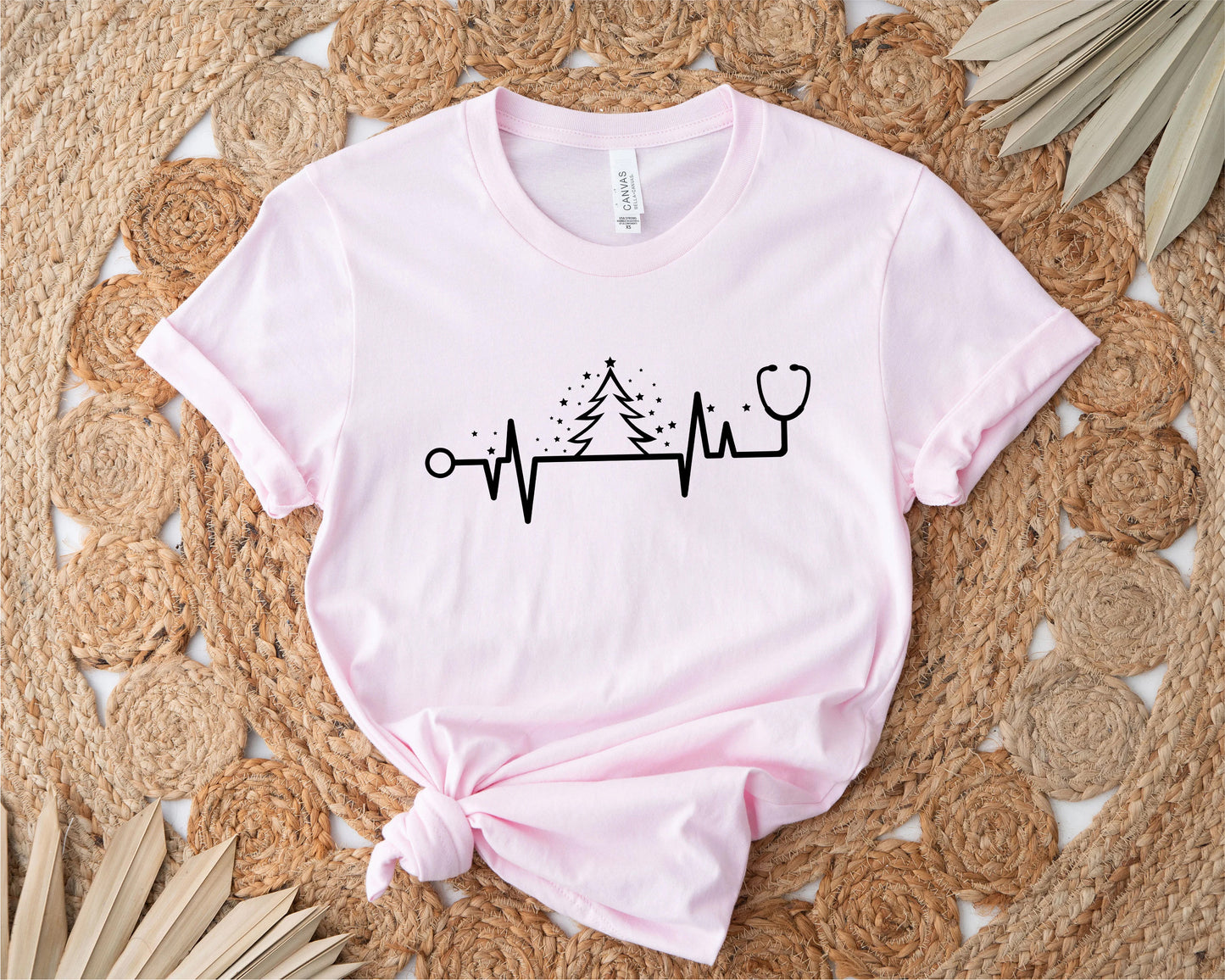 A pink t-shirt with a graphic design featuring a stethoscope and a Christmas tree, along with a line graph or line drawing, all placed on a woven mat.