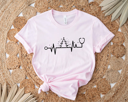 A pink t-shirt with a graphic design featuring a stethoscope and a Christmas tree, along with a line graph or line drawing, all placed on a woven mat.