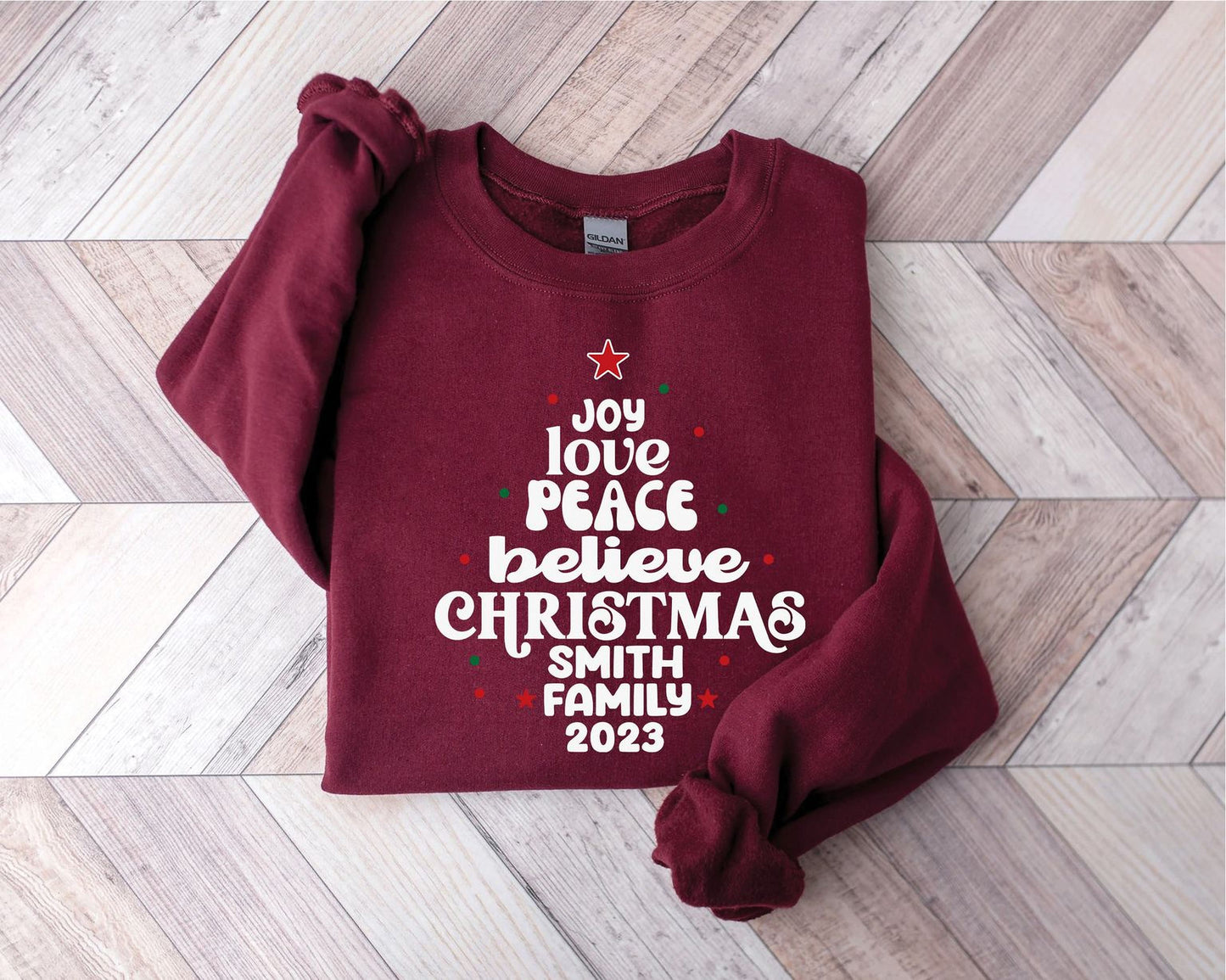 A burgundy sweatshirt with a Christmas-themed design printed on it, featuring the text "Joy, love, peace, believe, christmas, Smith, family, 2019" in a festive font.