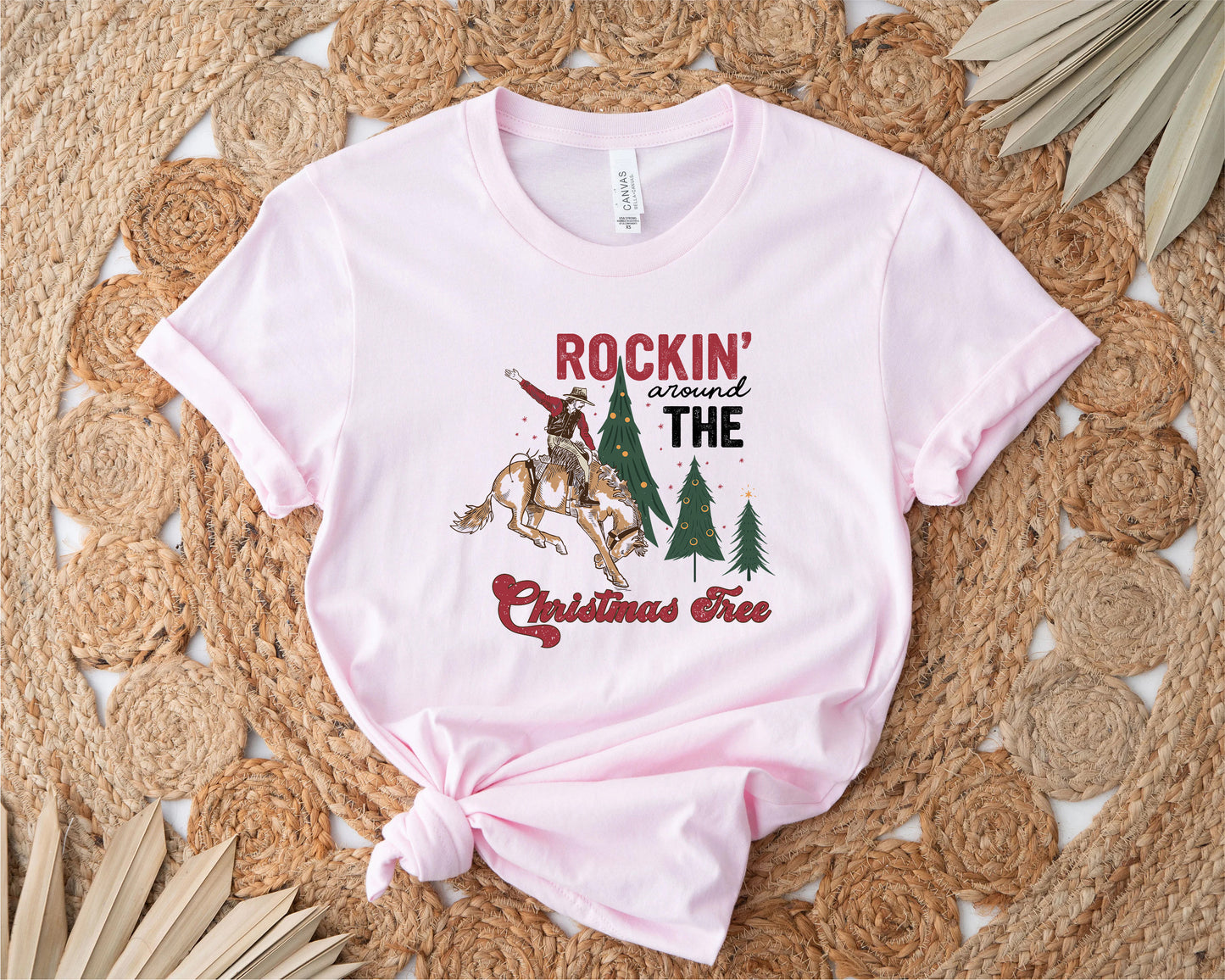 A pink t-shirt with a graphic design featuring a cowboy riding a bucking bronco, surrounded by Christmas trees, with the text "ROCKIN' around THE CHRISTMAS Tree" printed on it.