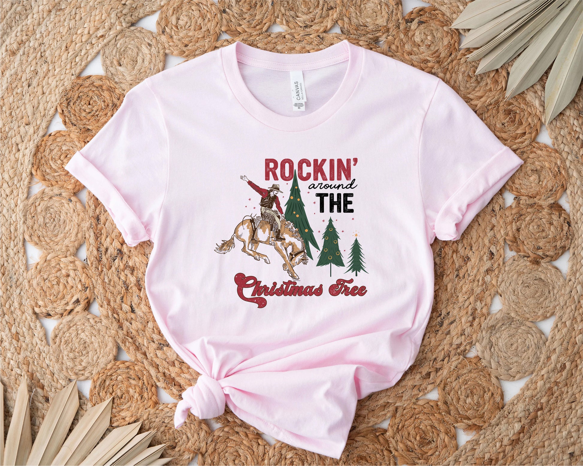 A pink t-shirt with a graphic design featuring a cowboy riding a bucking bronco, surrounded by Christmas trees, with the text "ROCKIN' around THE CHRISTMAS Tree" printed on it.