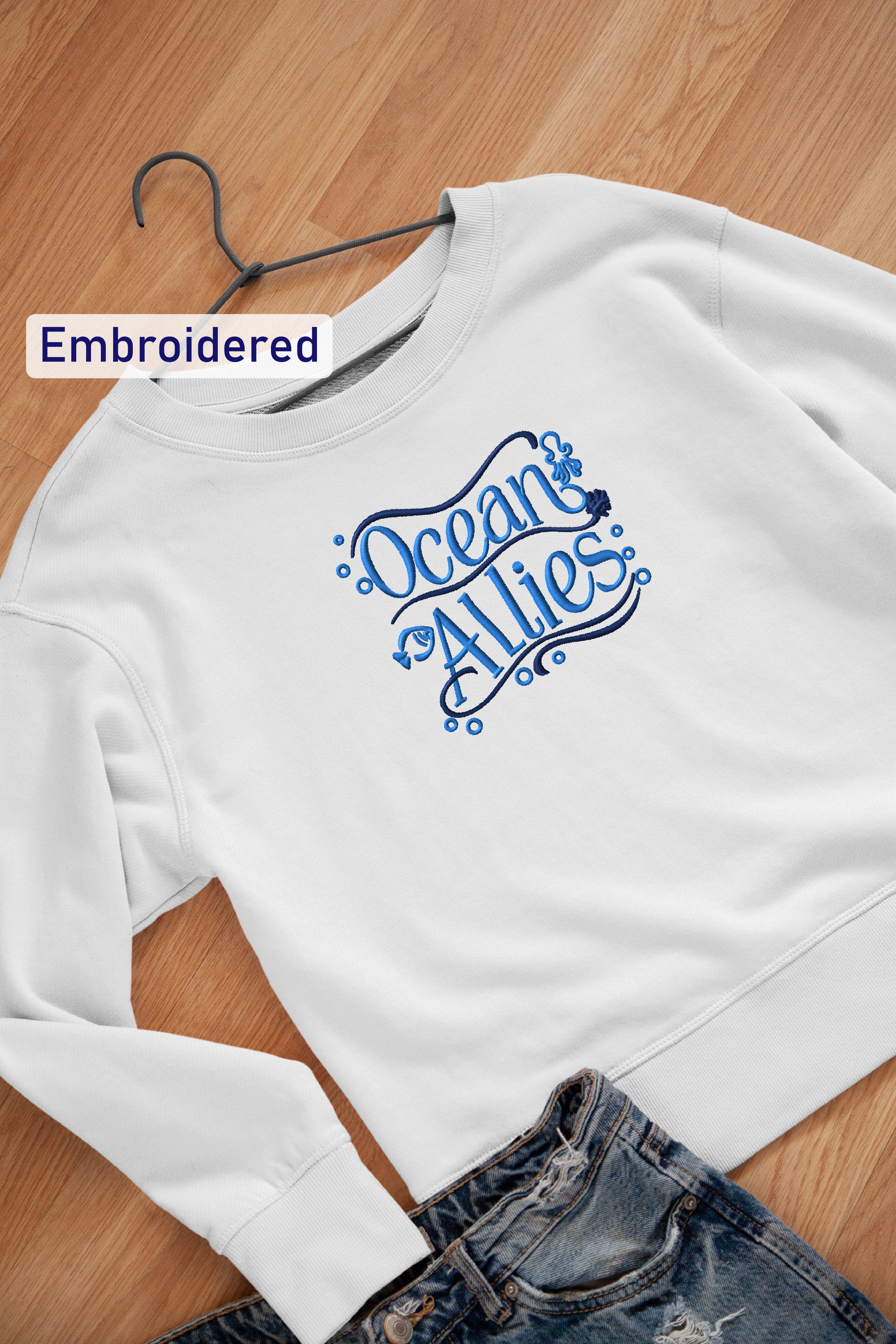 A white sweatshirt with the text "Ocean Allies" embroidered on it, along with a pair of blue jeans, are laid out on a wooden surface.