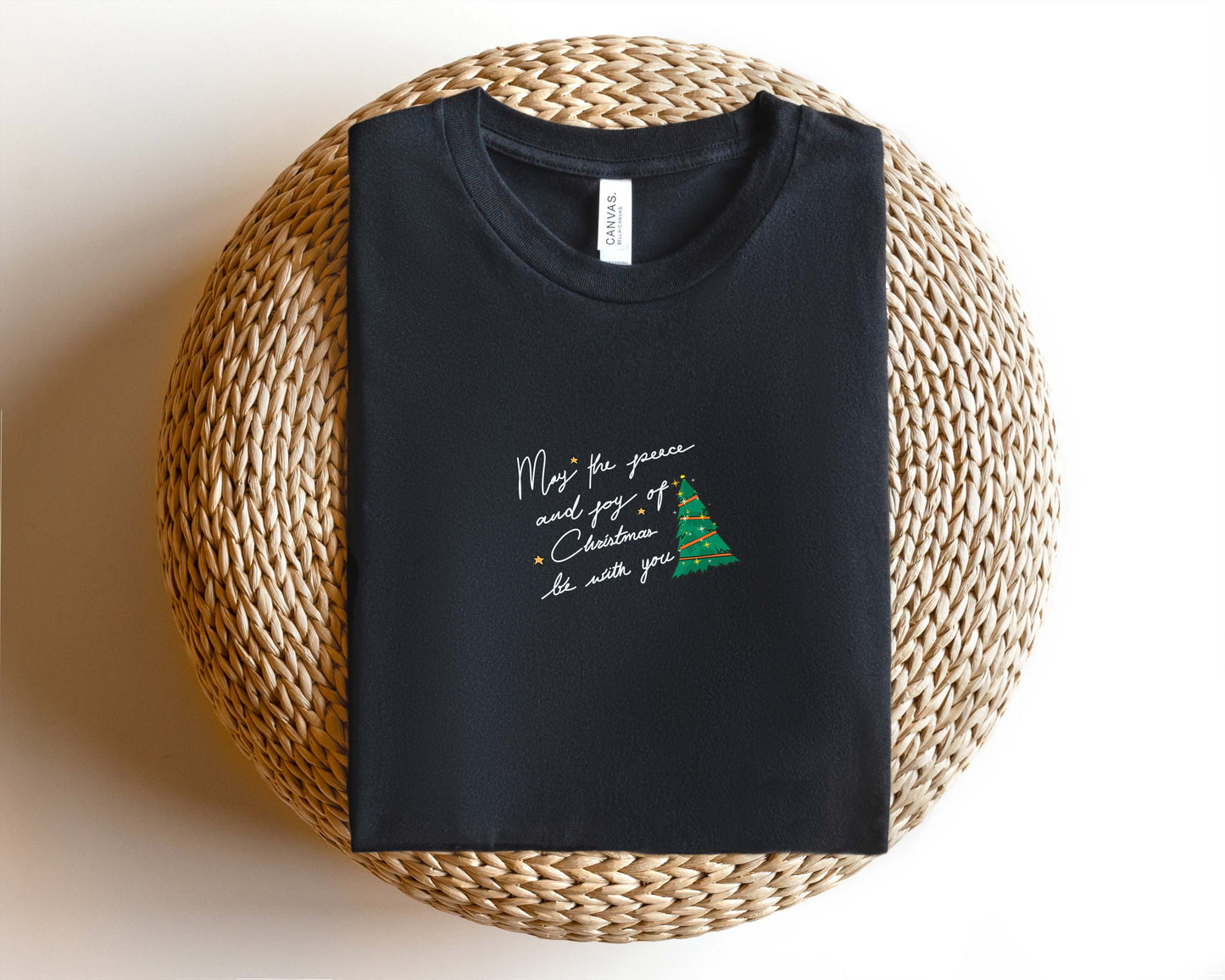 A black t-shirt with a Christmas tree design and the text "May the joy of Christmas be with you" printed on it, placed on a woven basket.