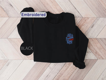 A black sweater with a blue embroidered design on the front, laid out on a wooden surface.