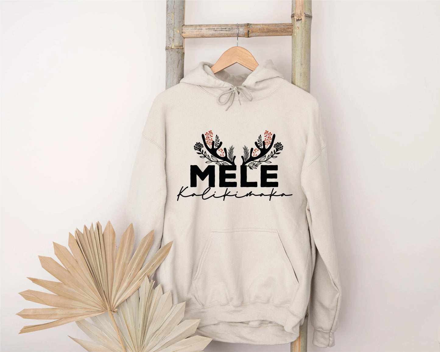 A beige hoodie with the text "MELE KALIKIMAKA" printed on it, hanging on a wooden ladder.