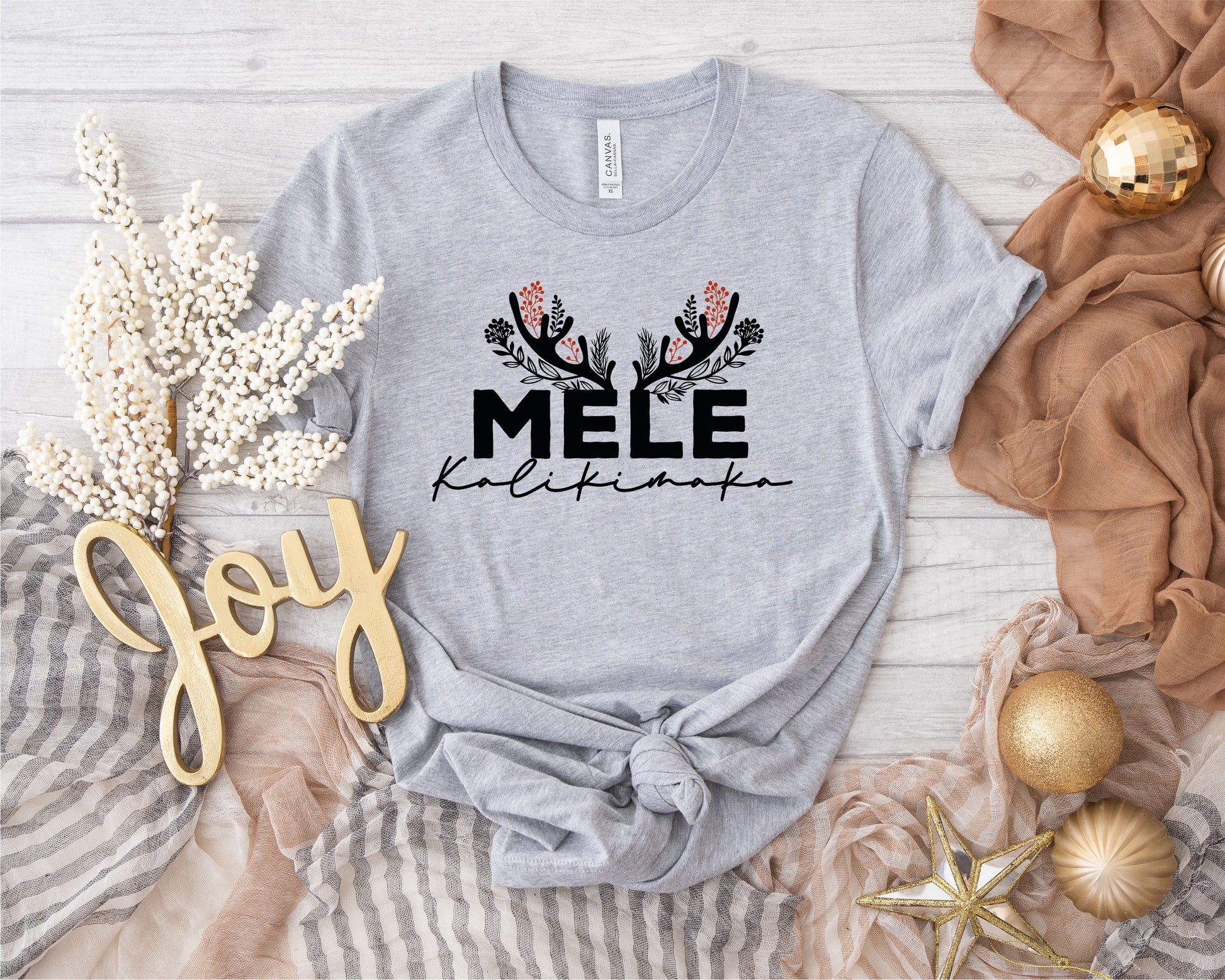 a gray t-shirt with a graphic design, surrounded by holiday-themed items such as a gold ornament, a white flower, and a gold star.