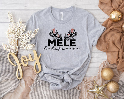 a gray t-shirt with a graphic design, surrounded by holiday-themed items such as a gold ornament, a white flower, and a gold star.
