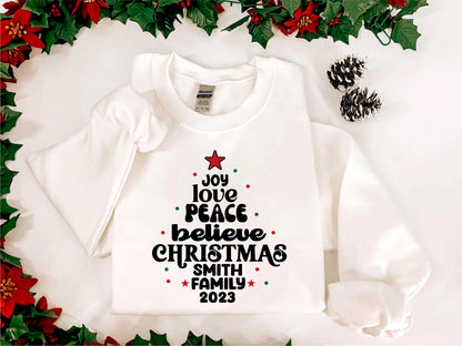 A white t-shirt with a Christmas tree design and text printed on it, surrounded by red poinsettia flowers and pine cones.