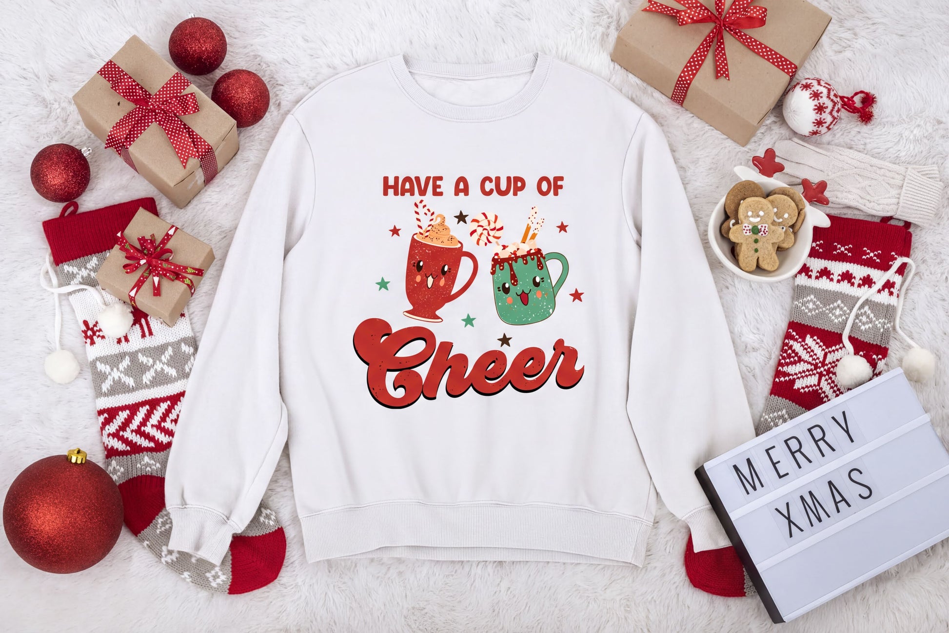 A white sweatshirt with a graphic design of two mugs and the text "Have a cup of cheer" is surrounded by various Christmas-themed items, including gift boxes, ornaments, and a "Merry Xmas" sign.