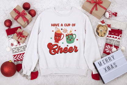 A white sweatshirt with a graphic design of two mugs and the text "Have a cup of cheer" is surrounded by various Christmas-themed items, including gift boxes, ornaments, and a "Merry Xmas" sign.