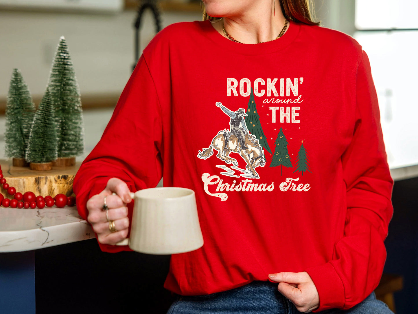 A person wearing a red sweater with a graphic design of a cowboy riding a bucking bronco, holding a mug, and surrounded by Christmas trees.