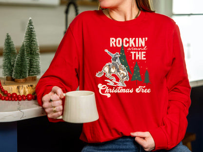 A person wearing a red sweater with a graphic design of a cowboy riding a bucking bronco, holding a mug, and surrounded by Christmas trees.