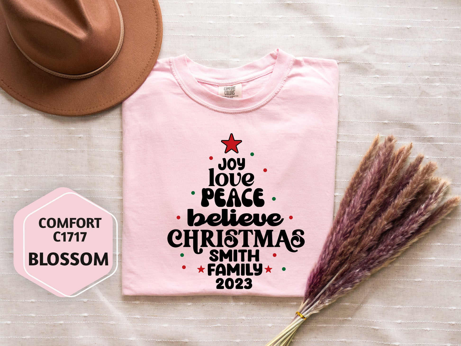 A pink t-shirt with a Christmas tree design and text, a brown hat, and some dried flowers are arranged on a white surface.
