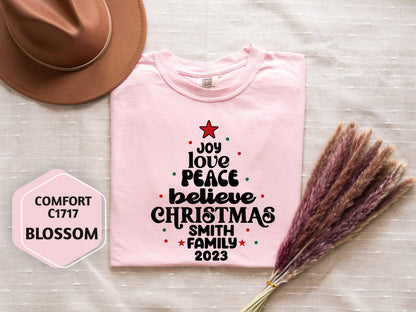 A pink t-shirt with a Christmas tree design and text, a brown hat, and some dried flowers are arranged on a white surface.