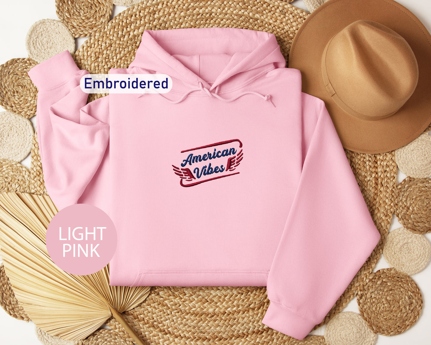 a pink hoodie with a red and blue embroidered logo, a brown hat, and a straw hat, all arranged on a woven mat.