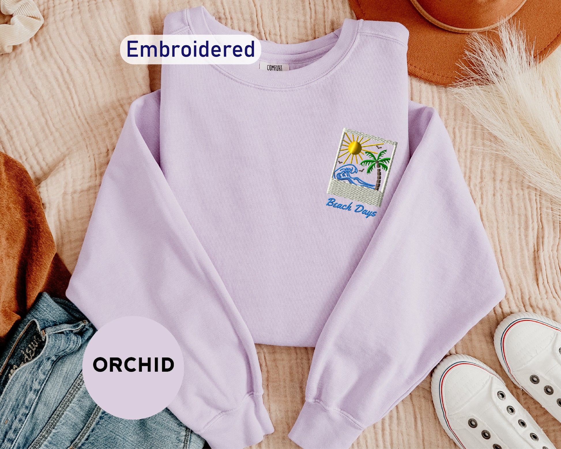 a purple sweater with a beach-themed embroidered patch, a pair of jeans, a hat, and a pair of white sneakers, all laid out on a surface.