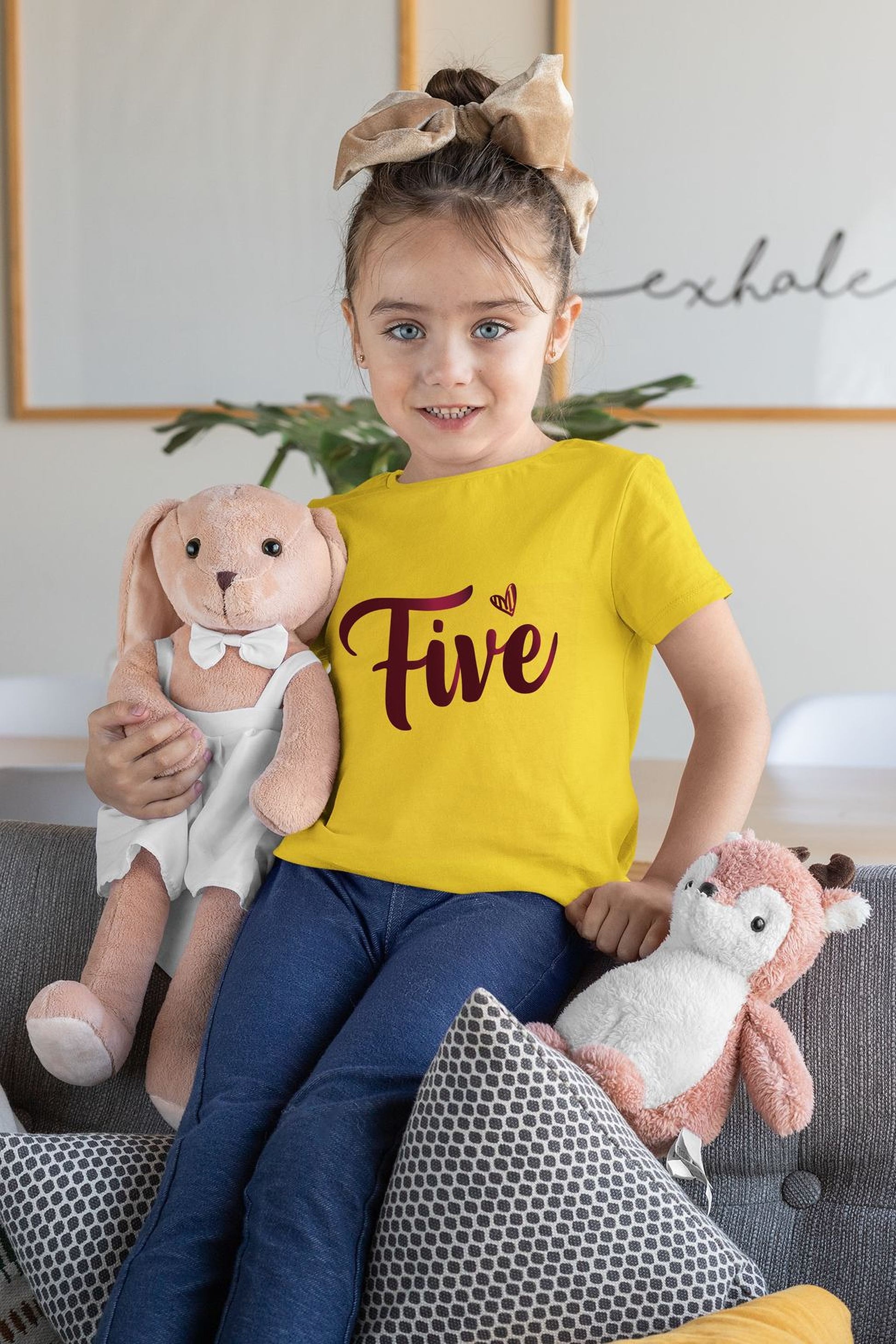 A young girl wearing a yellow t-shirt with the number five and holding a stuffed bunny rabbit and a stuffed deer.
