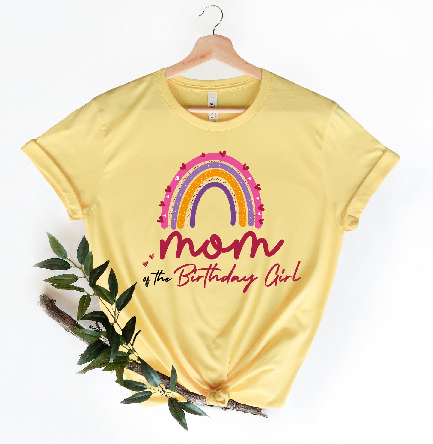 A yellow t-shirt with a colorful rainbow graphic and the text "MOM of the Birthday Girl" printed on it, hanging on a wooden hanger.