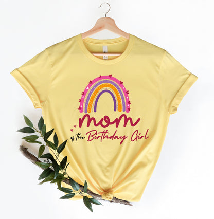 A yellow t-shirt with a colorful rainbow graphic and the text "MOM of the Birthday Girl" printed on it, hanging on a wooden hanger.