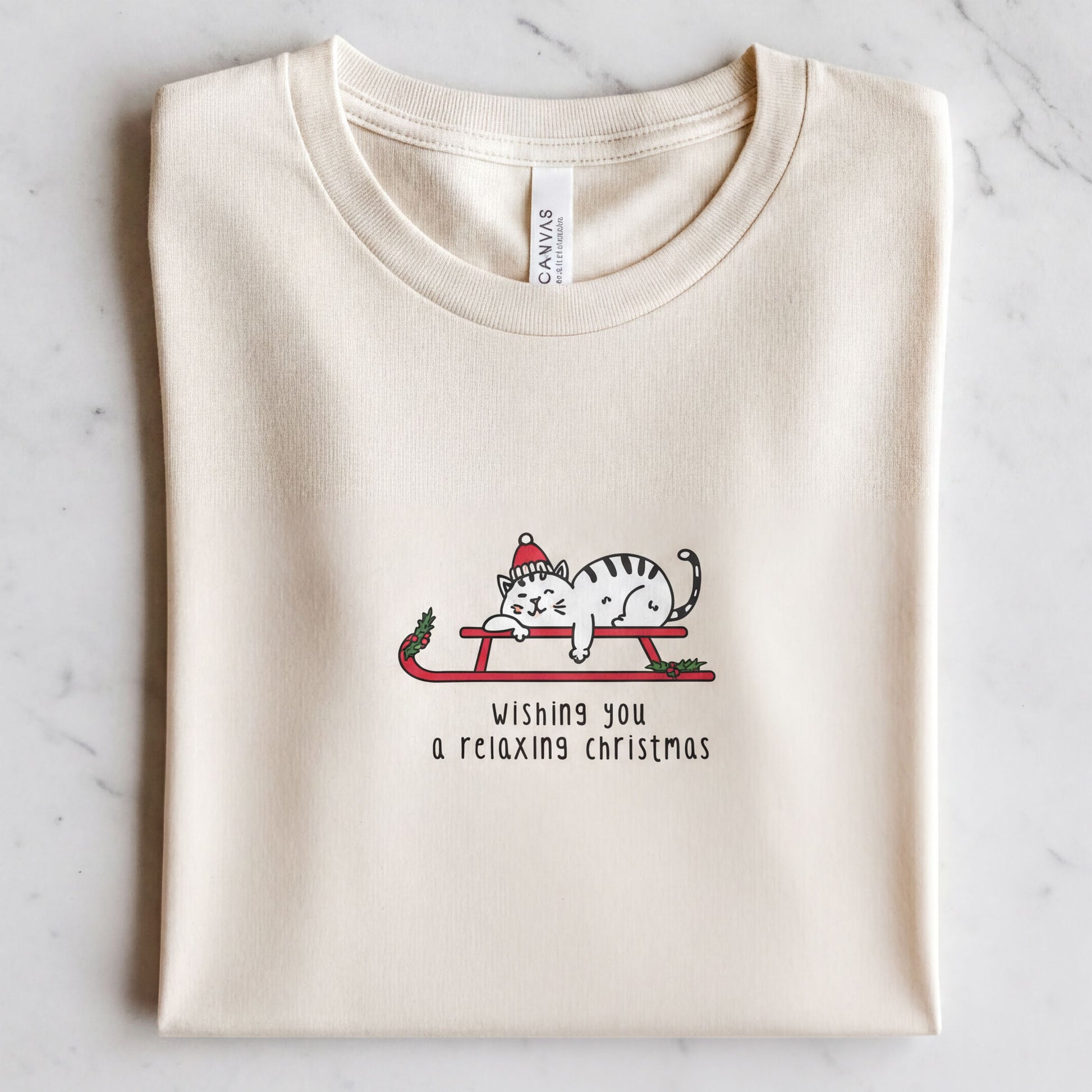 A white t-shirt with a graphic design depicting a cat wearing a Santa hat and riding a sled, along with the text "Wishing you a relaxing christmas".