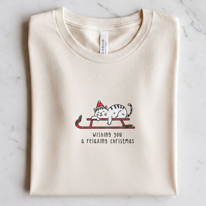A white t-shirt with a graphic design depicting a cat wearing a Santa hat and riding a sled, along with the text "Wishing you a relaxing christmas".