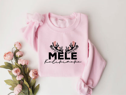 A pink sweatshirt with the text "MELE Kalikimaka" printed on it, along with a floral design. The sweatshirt is placed next to a bouquet of pink flowers.