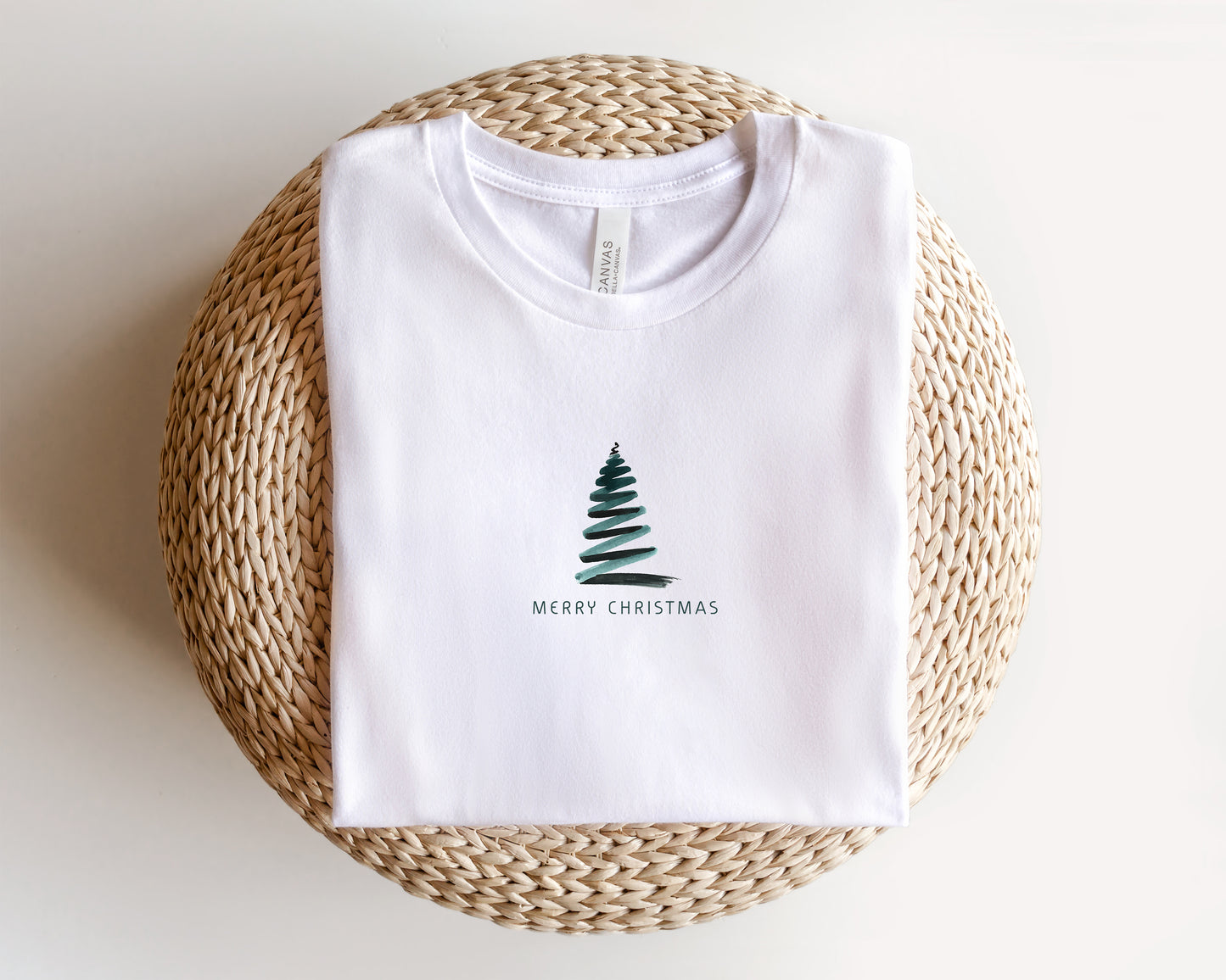 A white t-shirt with a Christmas tree design and the text "MERRY CHRISTMAS" printed on it, laid out on a woven basket.