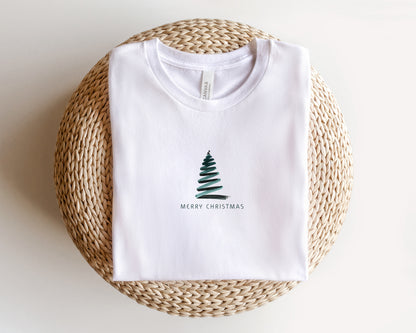 A white t-shirt with a Christmas tree design and the text "MERRY CHRISTMAS" printed on it, laid out on a woven basket.