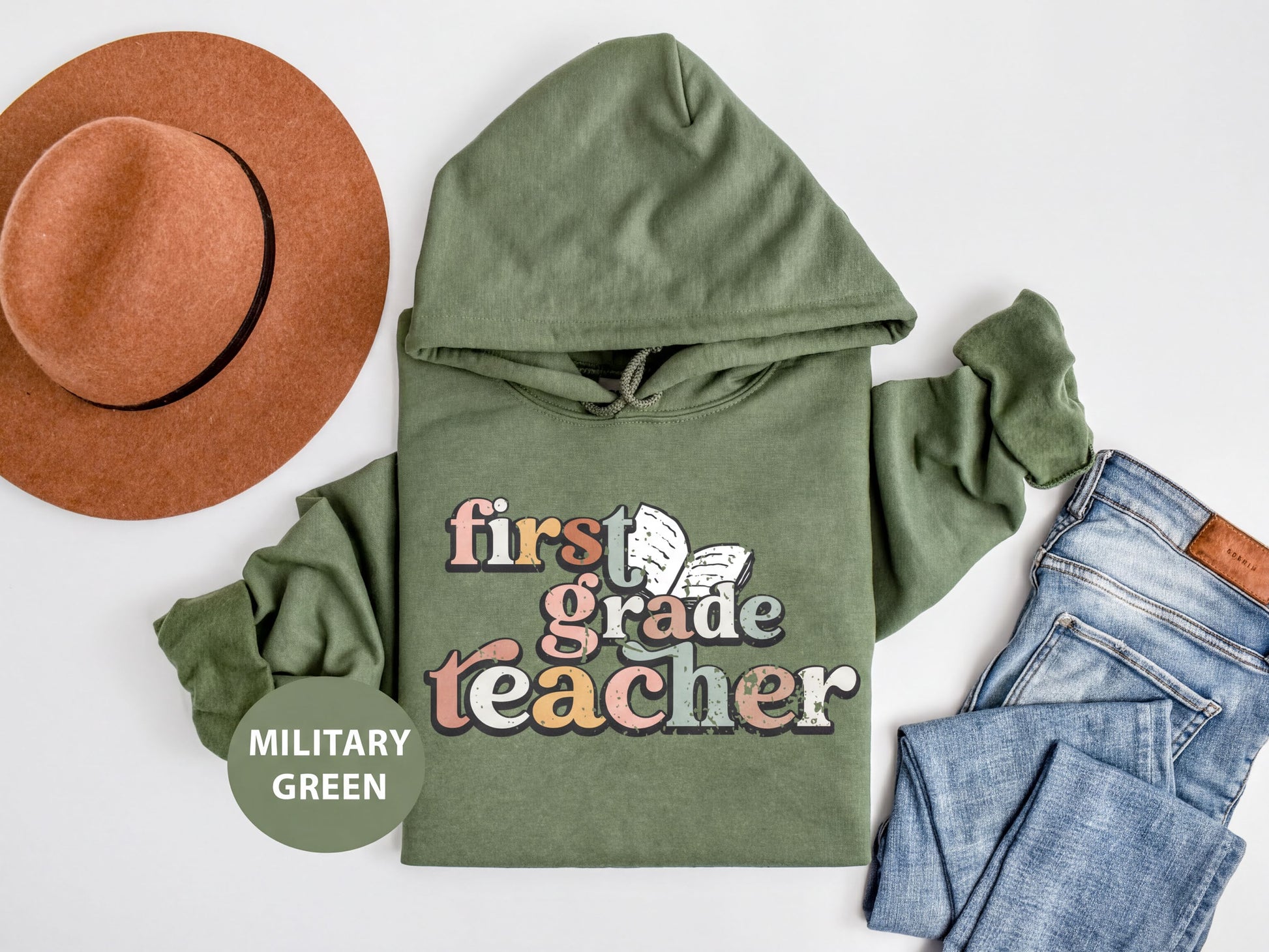 A military green hoodie with the text "first grade teacher" printed on it, along with a brown hat and a pair of blue jeans, all laid out on a white background.