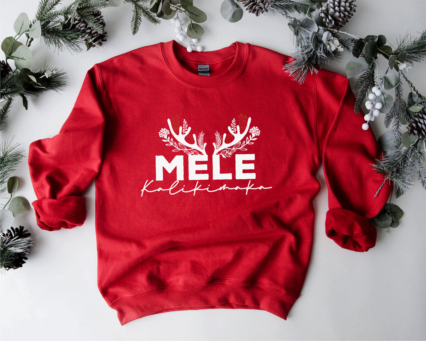 A red sweater with the text "MELE KOLIKINAS" printed on it, surrounded by pine branches and greenery.